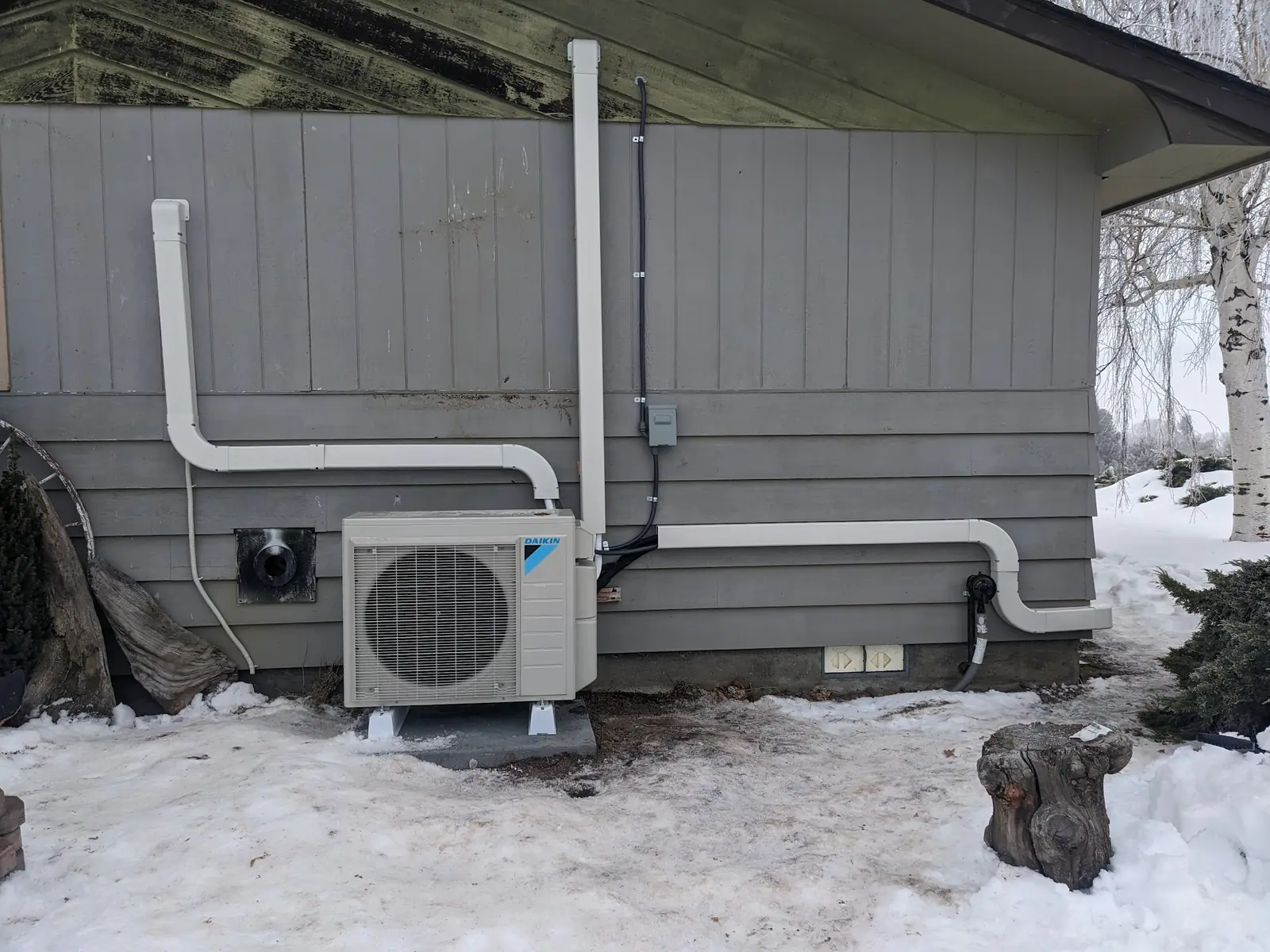 Daikin ductless outdoor unit with white line-set covers on grey siding, snow on the ground, installed by Total Quality Air